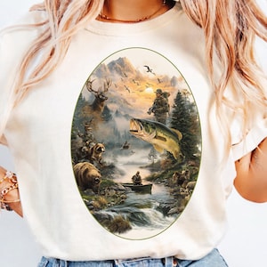 Puede incluir: Camiseta color crema con un gráfico ovalado de un paisaje montañoso con fauna. El gráfico incluye un ciervo, un oso, un pez y una persona en un bote. El fondo muestra una puesta de sol y pájaros.