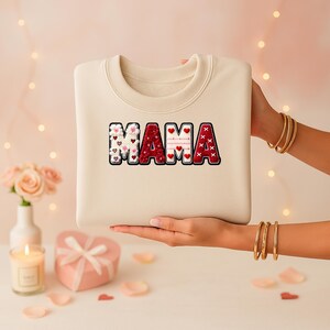 Op de afbeelding: Cr&egrave;mekleurige sweatshirt met het woord "MAMA" in een decoratief, meervoudig patroon. De letters hebben harten en andere patronen in rood, wit en roze. De sweatshirt wordt door een persoon vastgehouden.