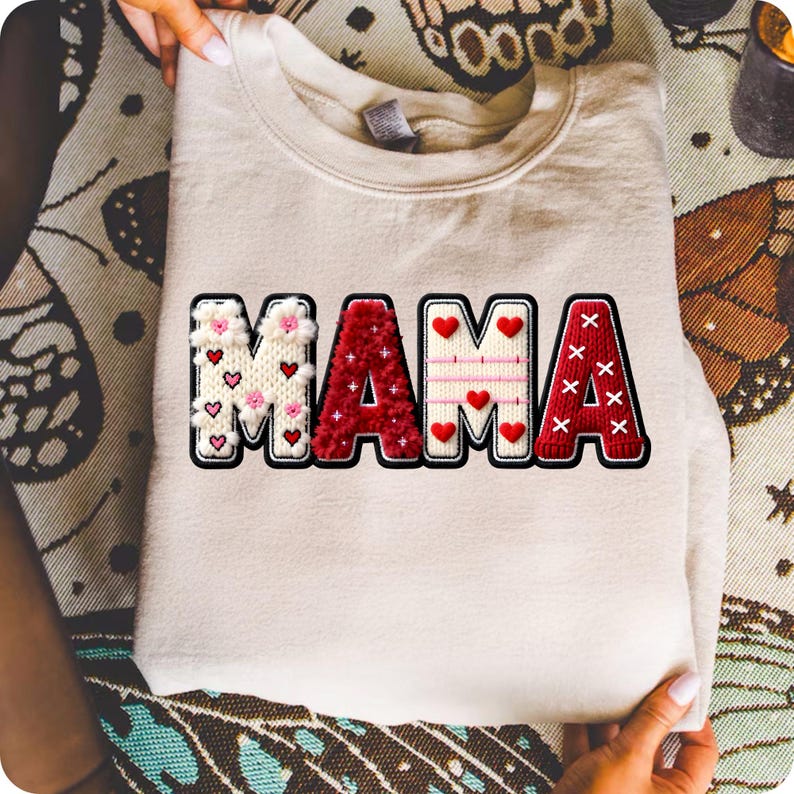 Op de afbeelding: Cr&egrave;mekleurige sweatshirt met het woord "MAMA" in een decoratief, gestructureerd ontwerp. De letters zijn zwart omlijnd en voorzien van rode harten en bloemaccenten, wat een gezellige en feestelijke uitstraling cre&euml;ert.