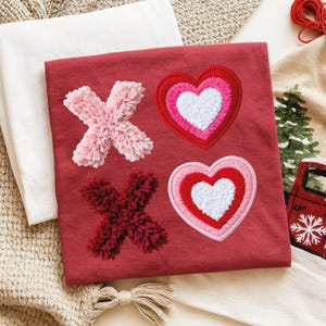 May include: A red t-shirt featuring embroidered X's and hearts. The X's are made of fluffy pink and burgundy material. The hearts are layered with white, pink, and red stitching. The shirt is folded on a white surface.