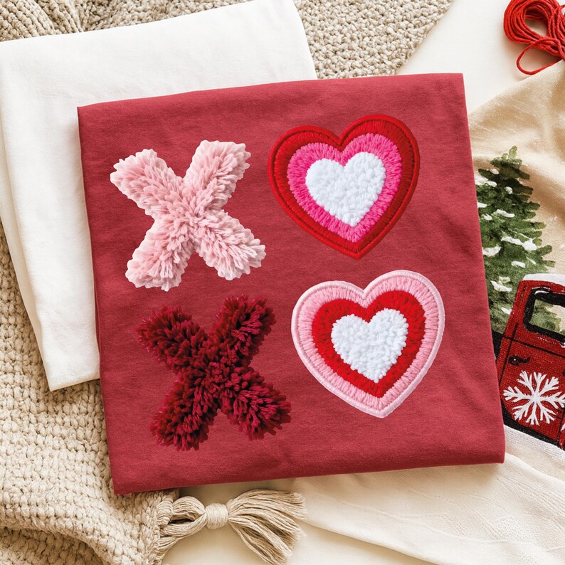 May include: A red t-shirt featuring embroidered X's and hearts. The X's are made of fluffy pink and burgundy material. The hearts are layered with white, pink, and red stitching. The shirt is folded on a white surface.