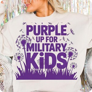 May include: Cream-colored sweatshirt with a purple graphic. The graphic features the text "PURPLE UP FOR MILITARY KIDS" with dandelion illustrations. The design includes purple dandelions and grass.