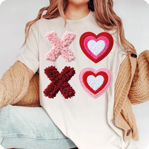 May include: Cream-colored t-shirt featuring four embroidered patches: a pink 'X', a burgundy 'X', and two hearts. One heart is red with a white center, and the other is pink with a white center. The patches are in a Valentine's Day theme.