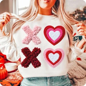 May include: White sweatshirt with pink and burgundy X's and two hearts. The hearts are red, pink, and white. The X's are made of a fuzzy material. The sweatshirt is a Valentine's Day theme.