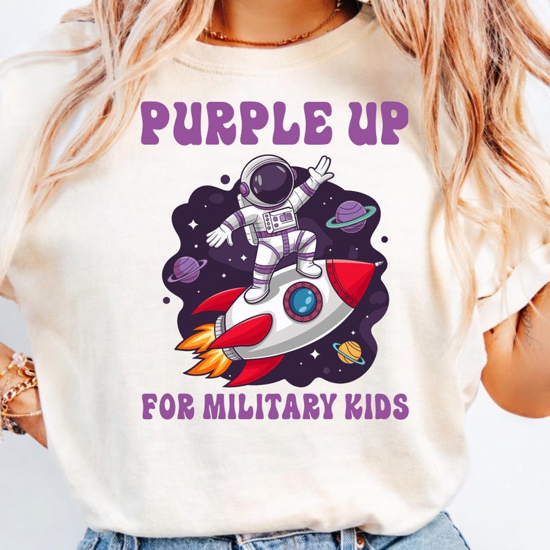 May include: Cream-colored t-shirt with the text "PURPLE UP" in purple, above an astronaut riding a red and white rocket ship. The phrase "FOR MILITARY KIDS" is printed below the rocket ship in purple.