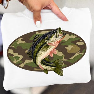 Pode incluir: Camisola branca com uma ilustração detalhada de um robalo saltando de um oval de camuflagem. O peixe é verde, preto e rosa, sobre um fundo de camuflagem castanho e verde.