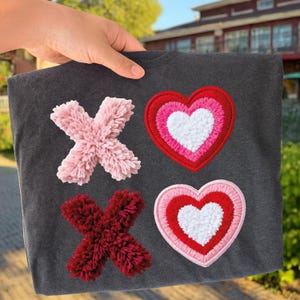 May include: Dark gray t-shirt featuring pink and burgundy pom-pom "X"s and two heart-shaped patches. The hearts have red, pink, and white layers. The shirt is held up by a hand.