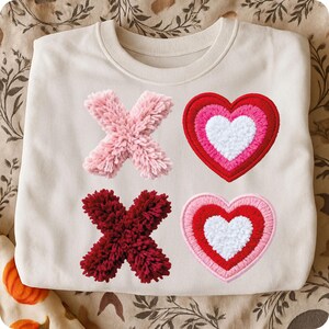 May include: Cream-colored sweatshirt with embroidered 'X' and heart designs. The 'X's are made of fluffy pink and burgundy material. The hearts are layered with red, pink, and white, creating a Valentine's Day theme.