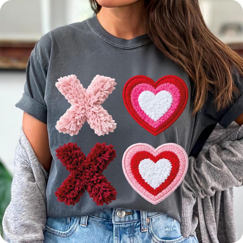 May include: Dark gray t-shirt featuring the letters "X" and hearts. The "X" letters are made of pink and red fluffy material. The hearts are red, pink, and white with a fluffy texture. The shirt is a casual style.