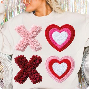 May include: Cream-colored sweatshirt featuring two fluffy "X" shapes, one pink and one burgundy, and two heart-shaped patches. The hearts are layered with red, pink, and white, creating a Valentine's Day theme. The design is embroidered.