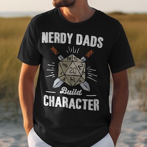Könnte beinhalten: Schwarzes T-Shirt mit einer weißen Grafik eines zwanzigseitigen Würfels mit Schwertern und dem Text "Nerdy Dads Build Character".