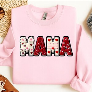 Op de afbeelding: Een lichtroze sweatshirt met het woord "MAMA" in een decoratief ontwerp. De letters zijn wit, rood en roze, met hart- en bloemaccenten. De sweatshirt heeft een ronde hals.