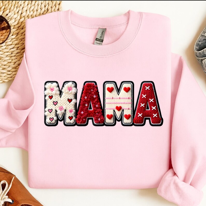 Op de afbeelding: Een lichtroze sweatshirt met het woord "MAMA" in een decoratief ontwerp. De letters zijn wit, rood en roze, met hart- en bloemaccenten. De sweatshirt heeft een ronde hals.