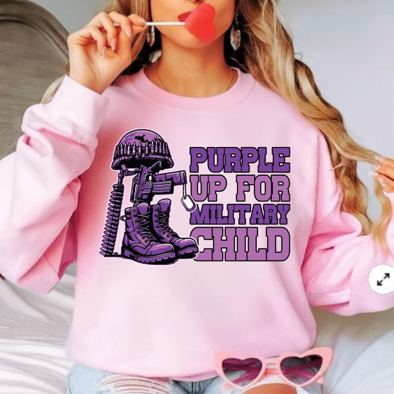 May include: Pink sweatshirt with a purple graphic. The graphic includes a military helmet, rifle, dog tags, and combat boots. Text on the sweatshirt reads "PURPLE UP FOR MILITARY CHILD".