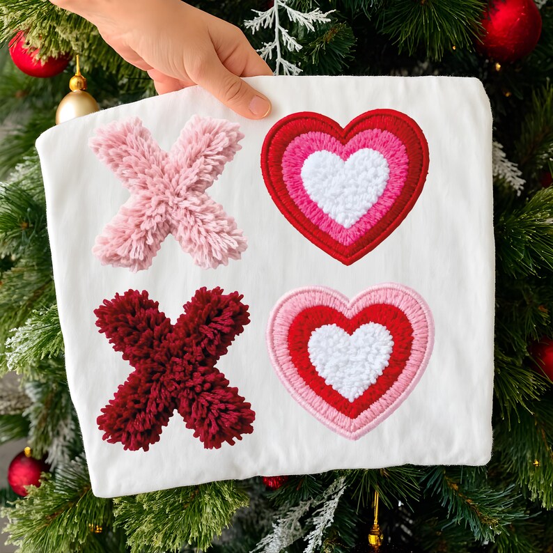 May include: A white fabric square featuring two "X" shapes and two heart shapes. The "X" shapes are made of fluffy pink and burgundy yarn. The hearts are red, pink, and white, with embroidered details. The item is held in front of a Christmas tree.