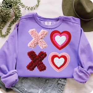 May include: A lilac-colored sweatshirt featuring the letters "X" and "O" in a pom-pom style, along with two heart designs. The hearts are in shades of red, pink, and white. The sweatshirt is made of a soft material.