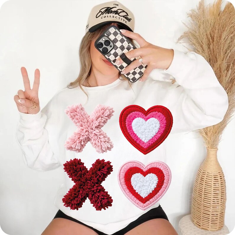 May include: White sweatshirt with pink and burgundy "X" and red and pink heart appliqu&eacute;s. The sweatshirt is a Valentine's Day theme. A tan baseball cap with the text "The Mark Collective" is worn.