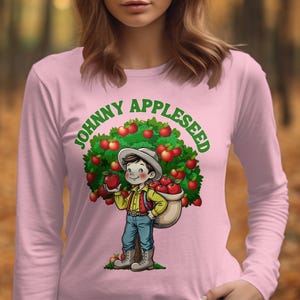 May include: Pink long-sleeve t-shirt with a cartoon illustration of Johnny Appleseed holding a basket of apples. The text "Johnny Appleseed" is written in a green and white font.