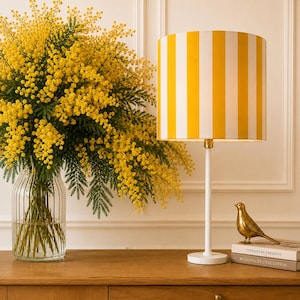 Peut inclure: Une lampe de table avec un abat-jour cylindrique à rayures verticales jaunes et blanches, un pied blanc fin et une base circulaire. À côté se trouvent une figurine d'oiseau dorée sur deux livres empilés et un vase en verre rempli de mimosa jaune.