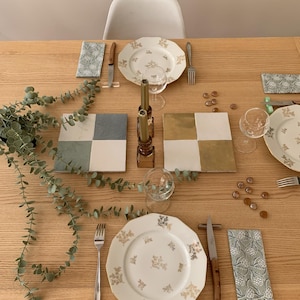 Peut inclure: Une table dressée avec deux couverts, chacun avec une assiette blanche à bordure dorée et un motif floral, une fourchette en argent et un couteau. Il y a deux jeux de chandeliers dorés, une plante verte et un jeu de carreaux dorés et blancs sur la table.