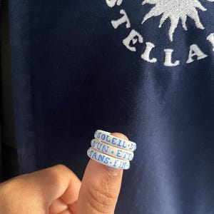 Puede incluir: Tres anillos de silicona blanca con letras azules. Los anillos dicen "Soleil d'un Ete Sans Fin".
