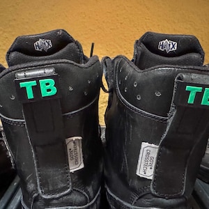 Könnte beinhalten: Ein Paar schwarze Lederstiefel mit grünen "TB"-Etiketten an den Seiten. Die Stiefel haben ein schwarz-weißes Etikett mit der Aufschrift "GORE-TEX CROSS TECH" an der Seite.