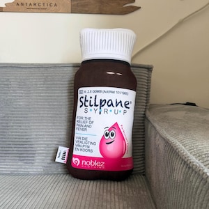 May include: A brown plush bottle of Stilpane syrup with a white cap and label. The label states "FOR THE RELIEF OF PAIN AND FEVER" and features a pink drop character. The bottle is on a gray corduroy surface.