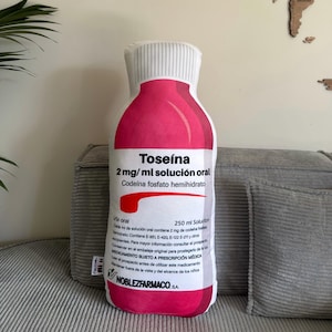 May include: A plush, oversized pillow shaped like a bottle of Toseína medicine. The pillow is primarily white and pink, with the medicine's name and dosage information printed on the front. The pillow is resting on a gray corduroy surface.