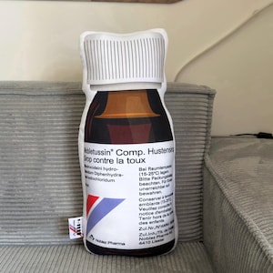 May include: A novelty cushion in the shape of a medicine bottle, with a white, lined cap and a brown liquid section. The label displays text in multiple languages, with red, blue, and white details. The cushion is placed on a grey corduroy surface.
