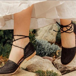 Op de afbeelding: Zwarte geweven platte sandalen met enkelbandjes. De sandalen hebben een spitse neus en een lage hak.