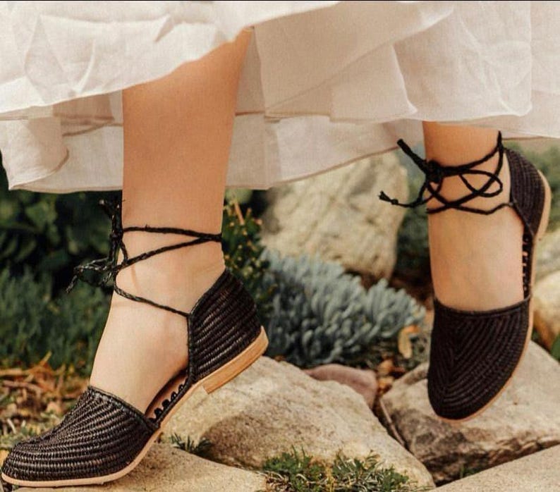 Op de afbeelding: Zwarte geweven platte sandalen met enkelbandjes. De sandalen hebben een spitse neus en een lage hak.