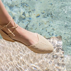 Op de afbeelding: Een paar beige gehaakte sandalen met een vetersluiting aan de enkel. De sandalen worden gedragen door iemand die in ondiep water staat.