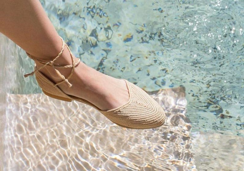 Op de afbeelding: Een paar beige gehaakte sandalen met een vetersluiting aan de enkel. De sandalen worden gedragen door iemand die in ondiep water staat.