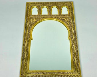May include: A gold-colored metal mirror with an ornate, intricate design. The mirror has a rectangular frame with a large arched opening in the center. The frame is decorated with a repeating pattern of small, intricate details.