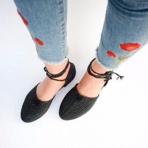 Op de afbeelding: Zwarte geweven platte schoenen met enkelbandjes. De schoenen hebben een kwastdetail op de teen.
