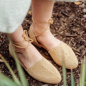 Op de afbeelding: Een paar beige geweven sandalen met enkelbandjes. De sandalen zijn gemaakt van een natuurlijk materiaal en hebben een eenvoudig, elegant ontwerp.