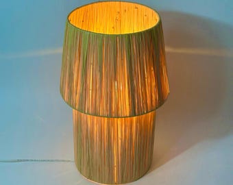 Puede incluir: Una lámpara de mesa con una pantalla tejida de fibras naturales. La pantalla es de color verde claro y tiene un brillo cálido y acogedor.