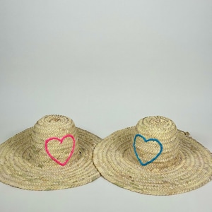 Pode incluir: Dois chapéus de sol de palha com abas largas. Um chapéu tem um coração rosa bordado, o outro um coração azul. Os chapéus são feitos de palha tecida, adequados para proteção solar.