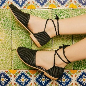 Op de afbeelding: Zwarte geweven platte sandalen met enkelbandjes. De sandalen staan op een betegelde vloer met een kleurrijk patroon.