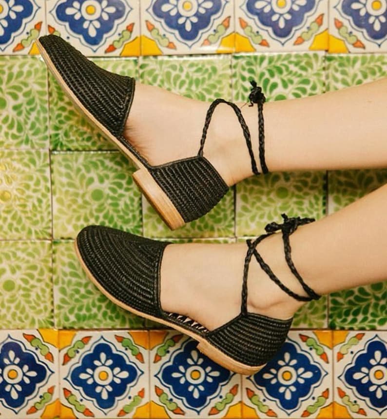 Op de afbeelding: Zwarte geweven platte sandalen met enkelbandjes. De sandalen staan op een betegelde vloer met een kleurrijk patroon.