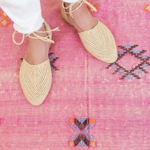 Op de afbeelding: Een paar beige geweven platte sandalen met enkelbandjes op een roze tapijt met patroon.