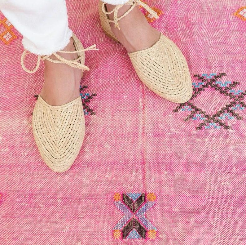 Op de afbeelding: Een paar beige geweven platte sandalen met enkelbandjes op een roze tapijt met patroon.
