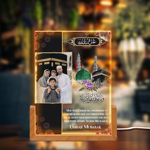 May include: A glowing glass photo frame with a family photo and Islamic calligraphy. The frame features a wooden base and a golden border. The text in the image reads "May your Umrah be a pilgrimage of immense blessings and forgiveness. May Allah accept your repentance and reward your every effort to seek His pleasure. Umrah Mubarak."