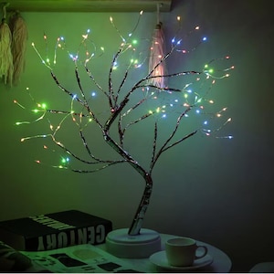 Könnte beinhalten: Eine dekorative LED-Lampe in Baumform mit silberfarbenem Stamm und Ästen. Die Äste sind mit kleinen, farbigen Lichtern geschmückt. Die Lampe steht auf einem weißen Sockel und steht auf einem Tisch neben einem Buch und einer Tasse.