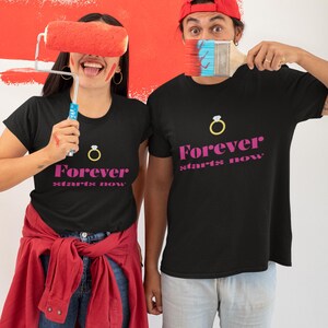 Op de afbeelding: Twee zwarte T-shirts met de tekst "Forever starts now" en een gouden ringafbeelding. De shirts worden gedragen door twee mensen die verfrollers vasthouden.