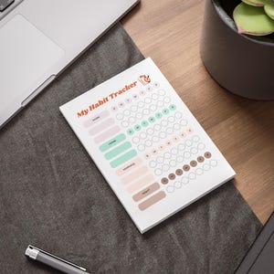 Könnte beinhalten: Weißer Notizblock zur Gewohnheitsverfolgung mit dem Titel "My Habit Tracker" in Orange. Der Notizblock hat Abschnitte für "Work", "Home", "Morning" und "Night" mit Kreisen zur täglichen Gewohnheitsverfolgung. Auf einer Holzoberfläche.