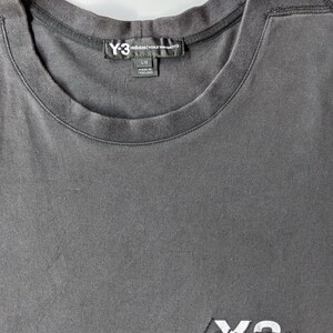 以下が含まれることがあります： ダークグレーのクルーネックTシャツ。右下に白で刺繍されたY-3 Adidasのロゴがあります。Y-3 Adidasのラベルが首元に見えます。シャツのサイズはLです。