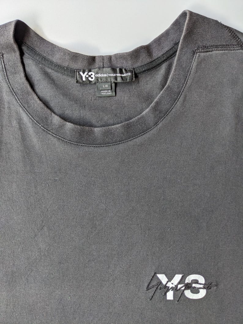 以下が含まれることがあります： ダークグレーのクルーネックTシャツ。右下に白で刺繍されたY-3 Adidasのロゴがあります。Y-3 Adidasのラベルが首元に見えます。シャツのサイズはLです。