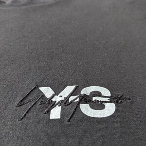以下が含まれることがあります： 白い「Y's」の刺繍ロゴが入った濃いグレーのTシャツのクローズアップ。 シャツはクルーネックで、柔らかく、質感のある生地で作られているようです。 ロゴは様式化されたフォントです。