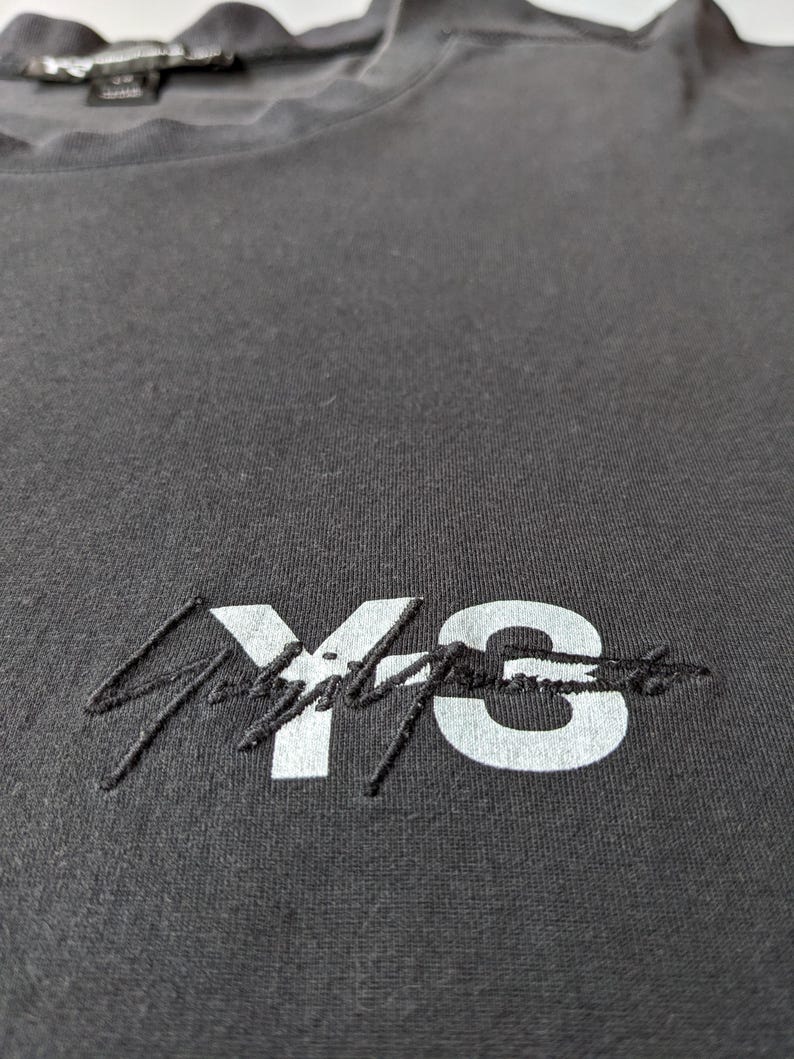 以下が含まれることがあります： 白い「Y's」の刺繍ロゴが入った濃いグレーのTシャツのクローズアップ。 シャツはクルーネックで、柔らかく、質感のある生地で作られているようです。 ロゴは様式化されたフォントです。